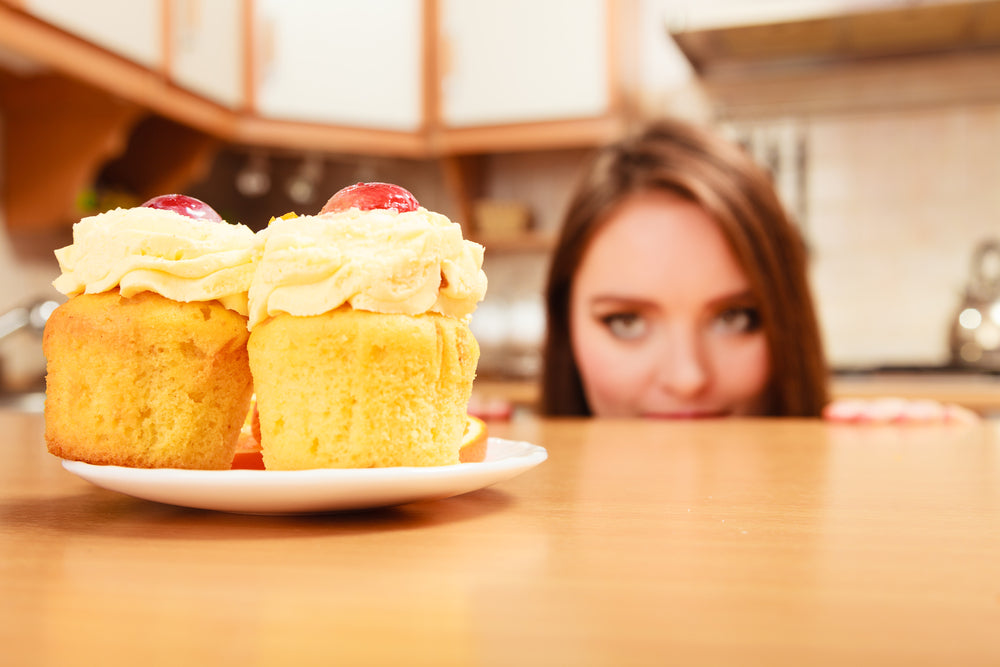 A woman with brown hair peeks over a kitchen counter, eyeing two frosted cupcakes with cherries on top, set on a white plate in a cozy kitchen setting.