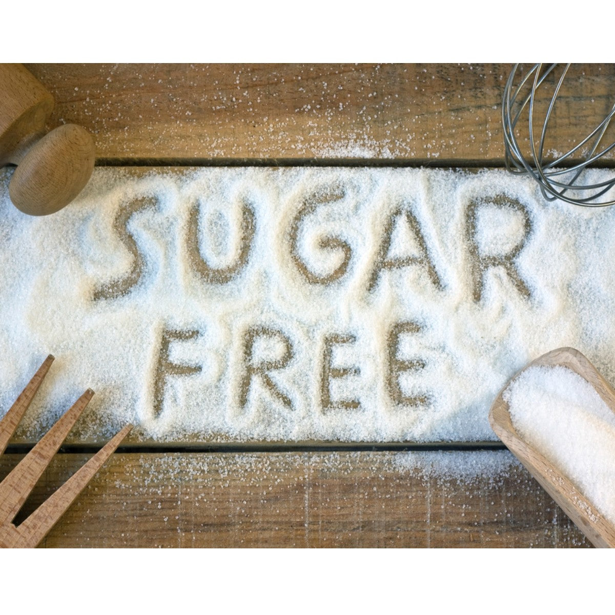 The words "SUGAR FREE" are written in brown sugar on a wooden surface, surrounded by a wooden spoon, a whisk, and measuring tools, emphasizing a healthy cooking theme.