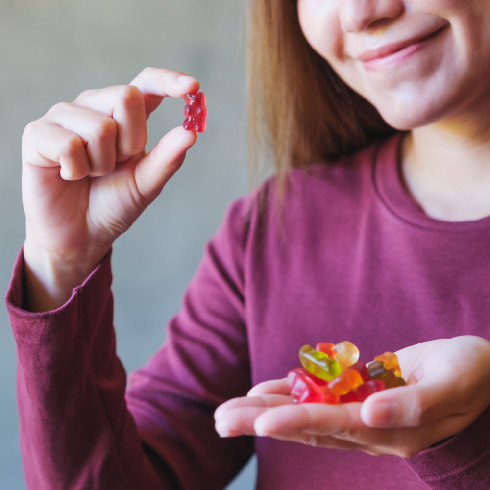 A smiling woman in a maroon shirt holds a gummy bear in one hand while displaying a handful of colorful sugar-free candies in the other, set against a neutral background.