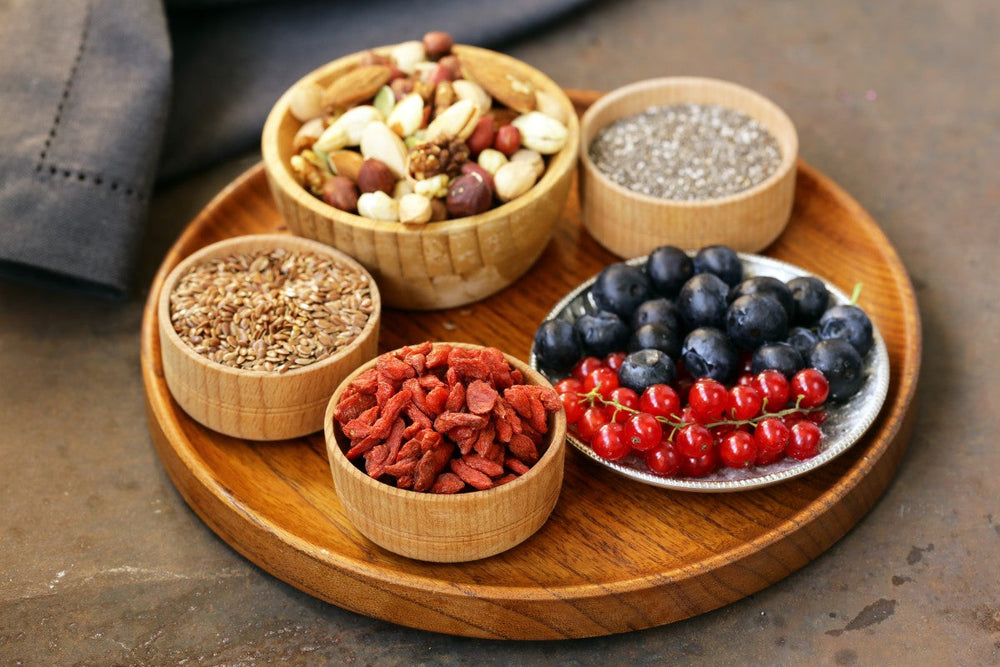 A wooden platter displays various superfoods, including bowls of goji berries, flaxseeds, and mixed nuts, alongside a silver dish of blueberries and red currants, set against a neutral background.
