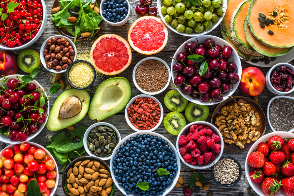 A vibrant assortment of fresh fruits, nuts, and seeds is arranged on a wooden surface, showcasing cherries, blueberries, avocados, and sliced melon among various bowls and scattered greenery.