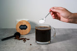 A hand holds a spoonful of sweetener above a clear mug of black coffee, with a container labeled "Stevia" and scattered coffee beans in the background on a gray countertop.