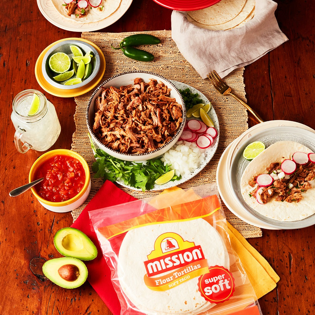A wooden table is set with a variety of taco ingredients, including pulled meat, tortillas, salsa, lime, jalapeños, and avocado, inviting a festive meal preparation.