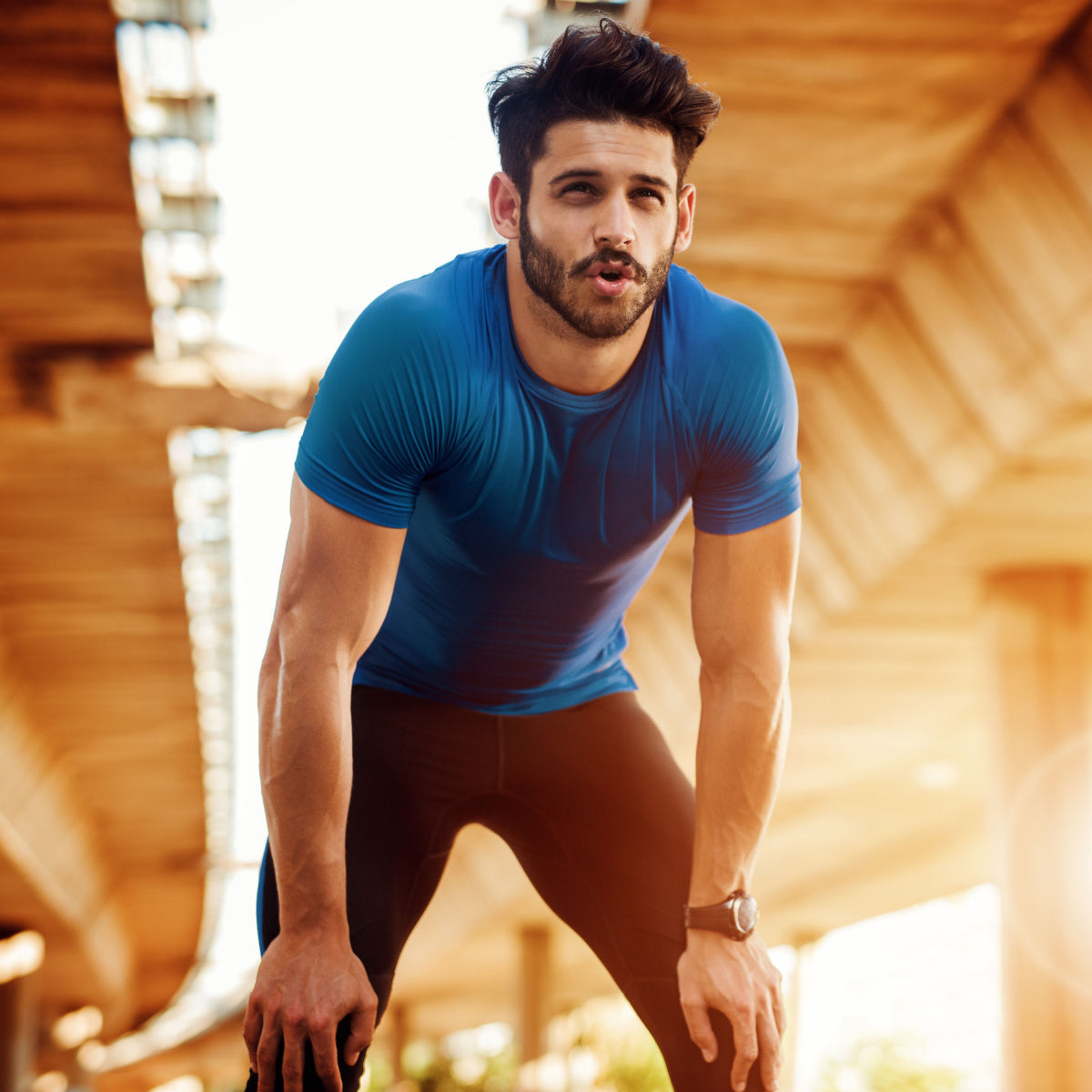 A man in a blue athletic shirt and black leggings leans forward, catching his breath, under a wooden structure in a bright outdoor setting.