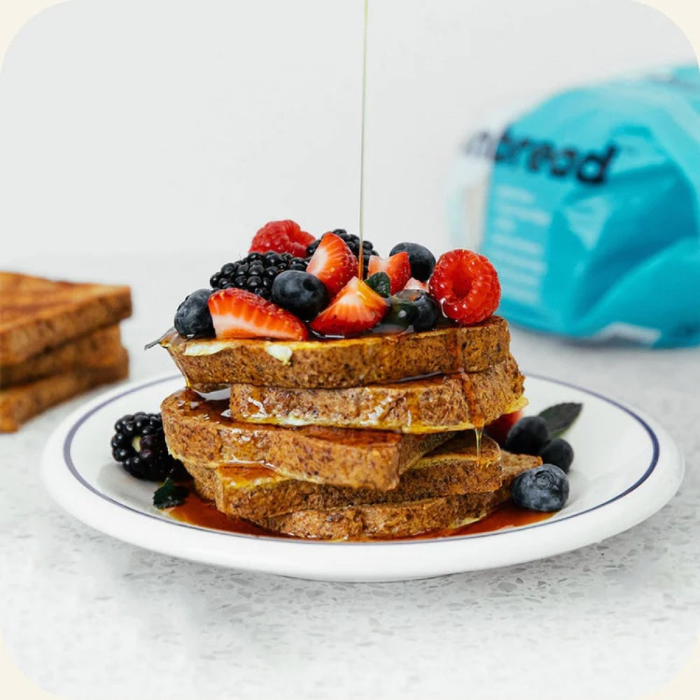 A stack of French toast topped with a variety of fresh berries and drizzled with syrup sits on a white plate against a light countertop, with a blue bag of bread visible in the background.