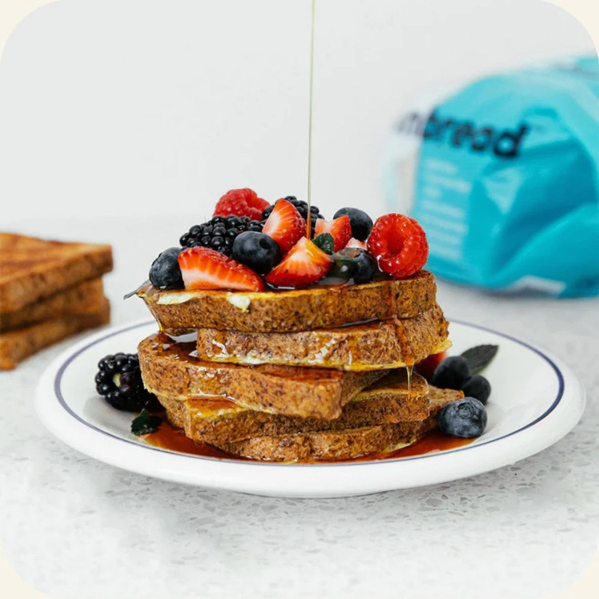 A stack of French toast topped with a variety of fresh berries and drizzled with syrup sits on a white plate against a light countertop, with a blue bag of bread visible in the background.