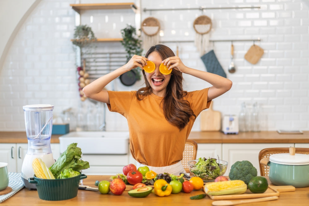 A woman in a brown shirt playfully holds orange slices in front of her eyes while smiling, surrounded by fresh vegetables and fruits on a kitchen counter, with a blender nearby.