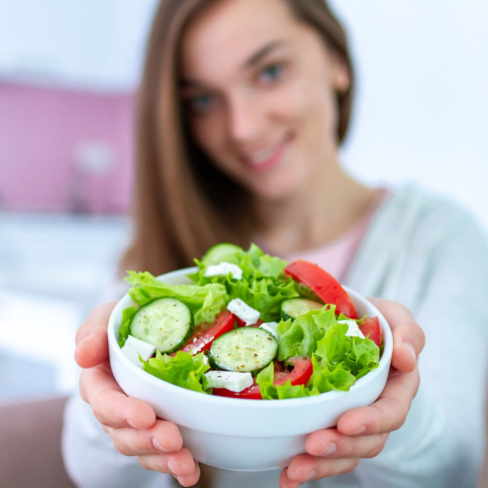 A smiling woman holds a bowl of fresh salad with lettuce, cucumbers, and tomatoes, showcasing the dish in a bright kitchen setting.