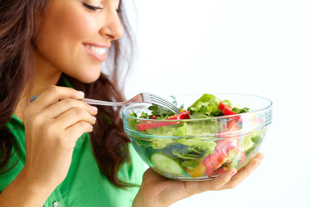 A woman in a green shirt smiles while holding a fork and a large bowl of fresh salad, emphasizing the importance of vegetables in a healthy diet.