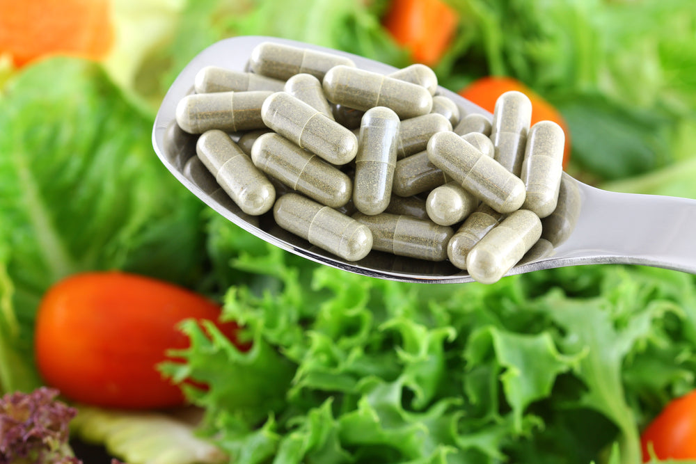 A silver spoon holds several green capsules above a vibrant mix of fresh vegetables, including leafy greens and tomatoes, highlighting vegan supplements for health.