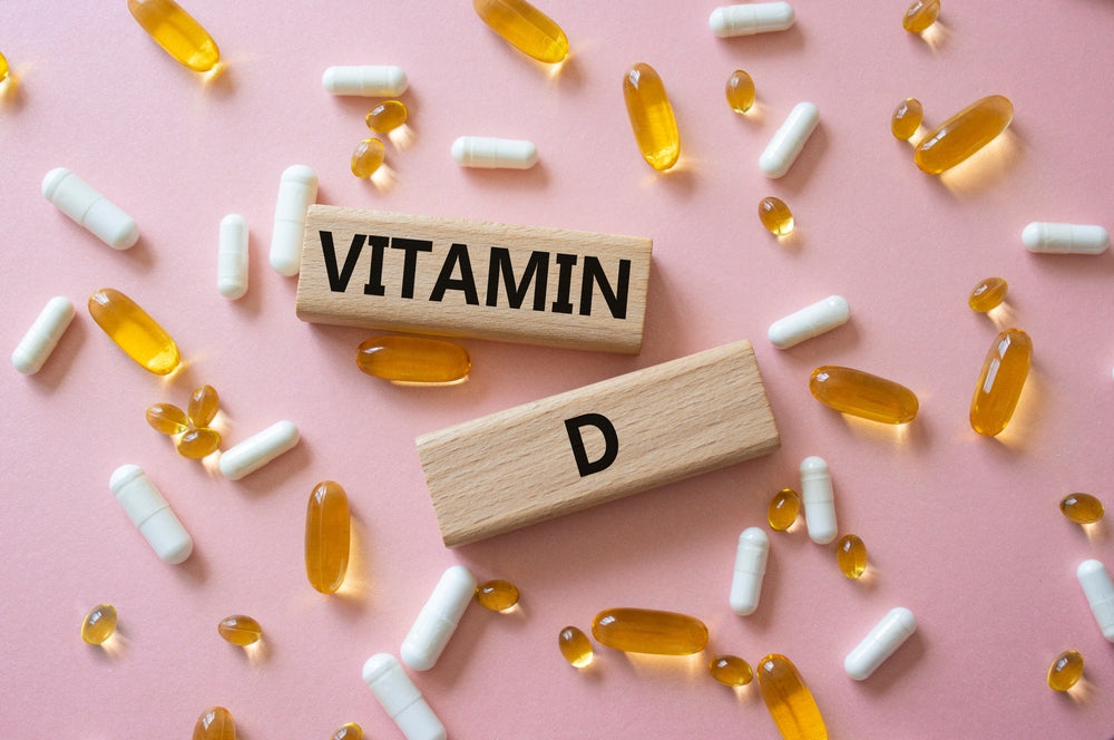 Wooden blocks spelling "VITAMIN" and "D" are surrounded by various yellow and white capsules on a pink background.