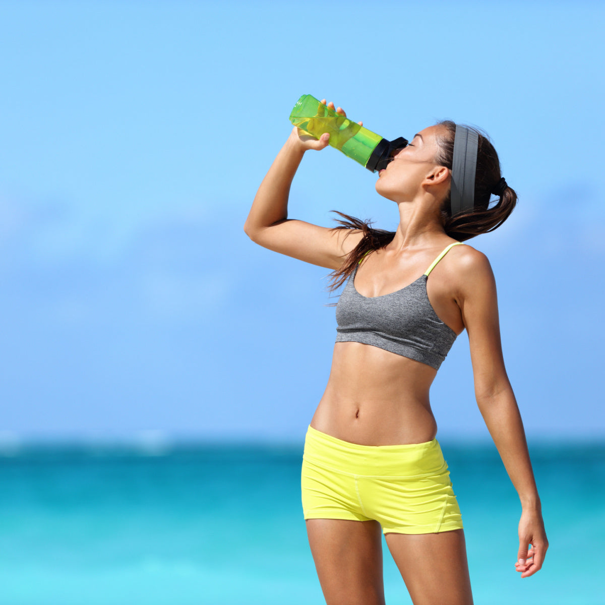 A young woman in a gray sports bra and yellow shorts drinks from a green water bottle while standing on a beach with turquoise water and a clear blue sky in the background.