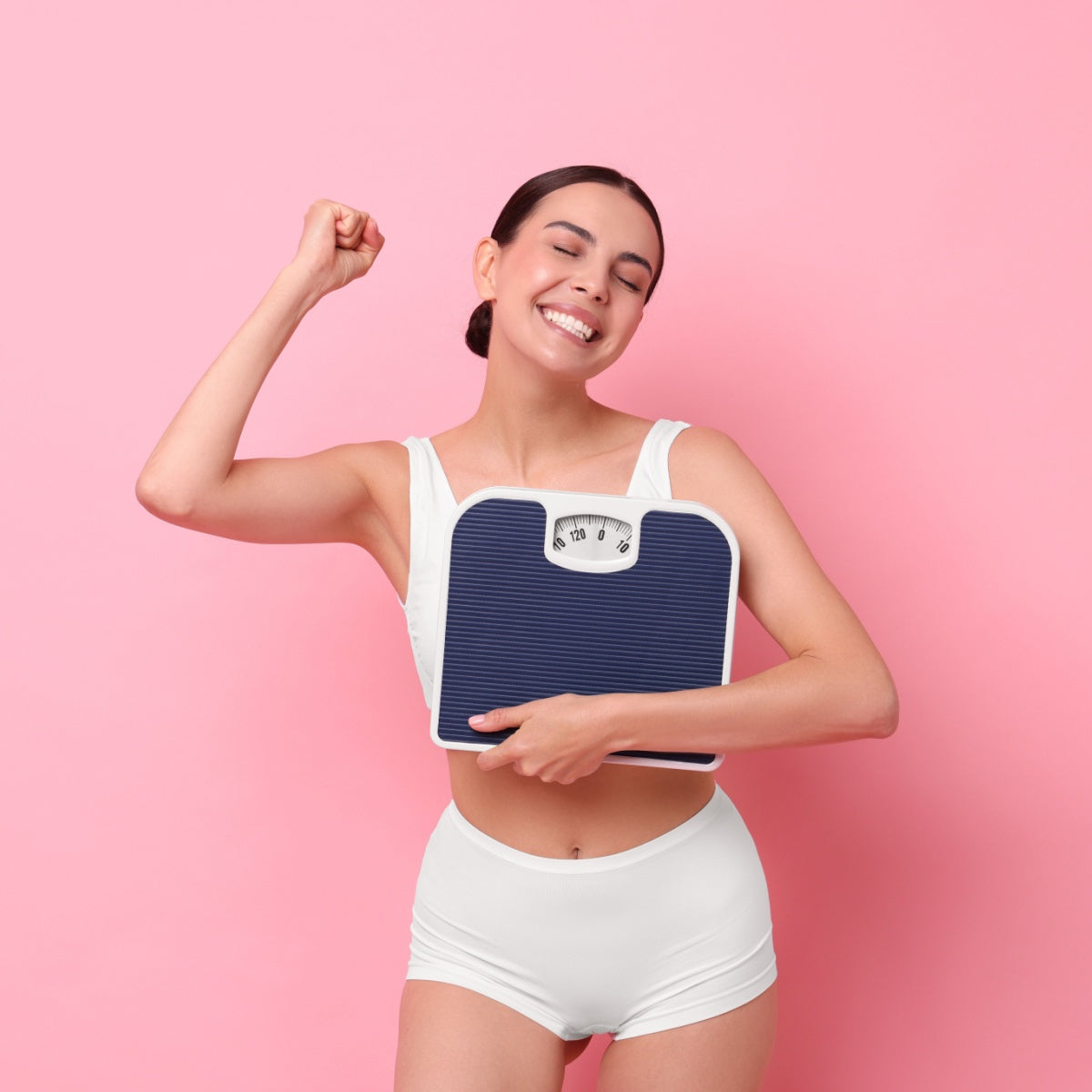 A young woman in a white outfit smiles broadly while holding a scale close to her chest, raising her fist in celebration against a pink background.