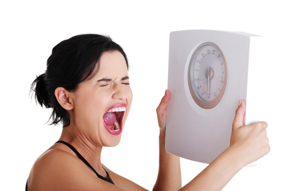 A woman with dark hair yells in frustration while holding a bathroom scale in front of her, expressing strong emotions in a bright, plain setting.