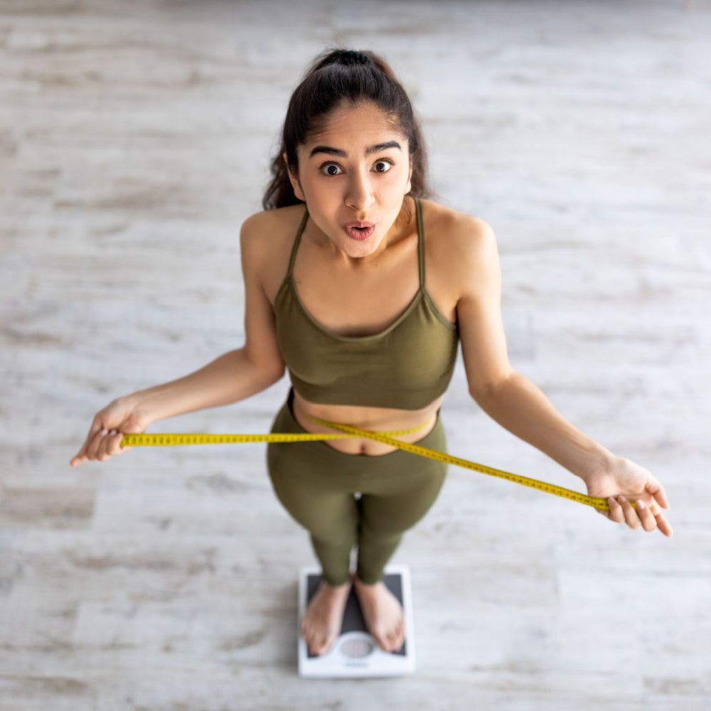 A surprised woman in a green workout outfit stands on a scale, holding a measuring tape around her waist, in a bright room with wooden flooring.