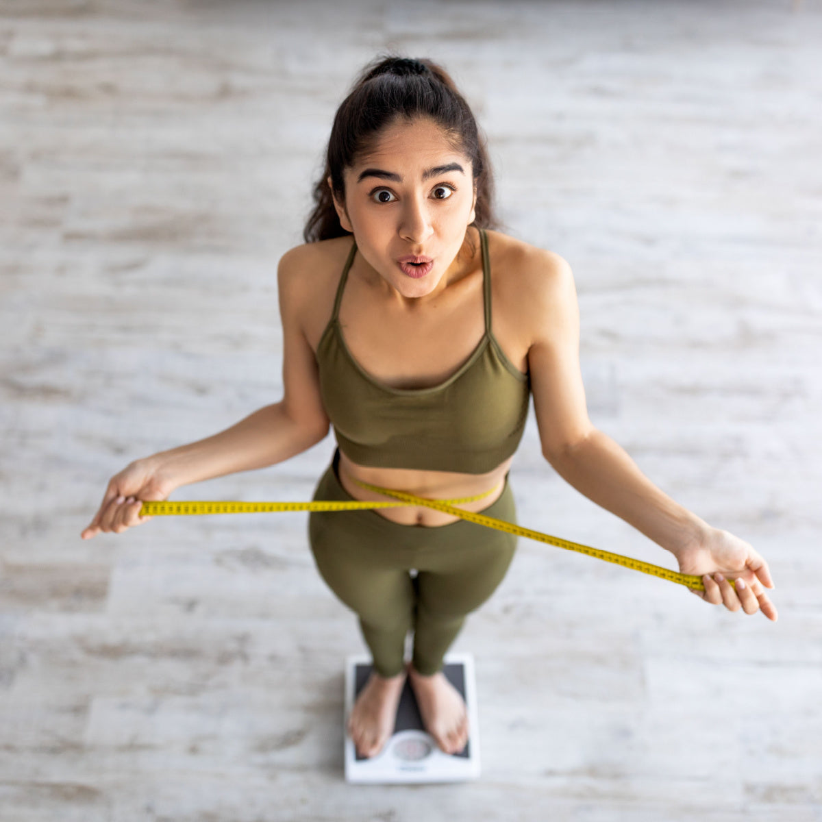 A surprised woman in a green workout outfit stands on a scale, holding a measuring tape around her waist, in a bright room with wooden flooring.