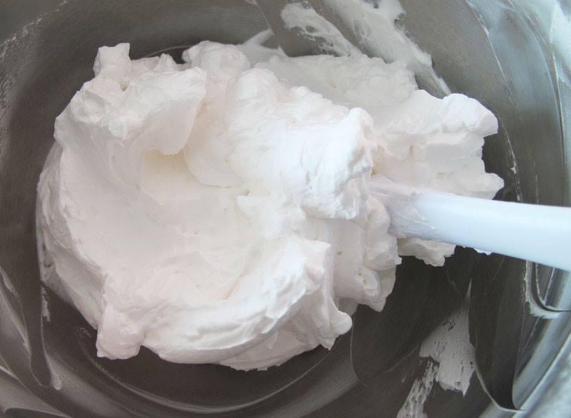 A mixing bowl contains fluffy, white whipped cream, with a spatula partially submerged, indicating preparation for a dessert or topping.