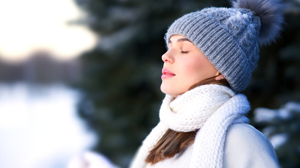 A young woman in a gray knitted hat and white scarf stands outdoors in a snowy setting, with her eyes closed and a serene expression, enjoying the fresh winter air.