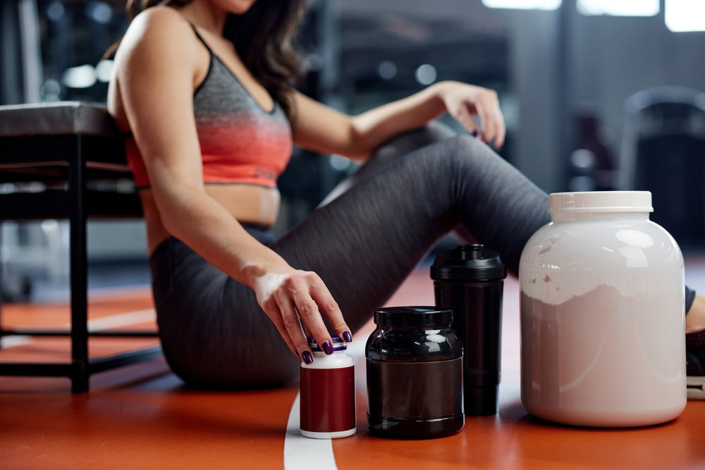 A person in workout attire sits on a gym floor, reaching for a supplement container while surrounded by various protein powders and shakes, emphasizing pre- and post-workout nutrition.