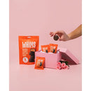 A hand is reaching for a dark chocolate peanut butter cup from a pink gift box filled with more cups and surrounded by roses, set against a soft pink background.