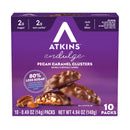 A purple box of Atkins Endulge Pecan Caramel Clusters displays the product name and nutritional information, with chocolate-covered clusters and pecans shown alongside the box.