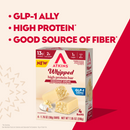 A box of Atkins Whipped high protein bars in Coconut Crème flavor, featuring a partially opened bar and text highlighting its protein content, low sugar, and fiber benefits against a colorful background.