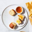 A round white plate holds three soft, golden-brown rolls, one cut open with butter and orange jam, alongside a small bowl of jam, set against a light marble background.