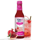 A bottle of Jordan's Skinny Mixes Grenadine syrup stands next to a glass filled with a sparkling drink, garnished with a cherry, alongside a fresh pomegranate cut in half.