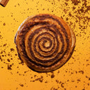 A freshly baked cinnamon roll with a golden-brown spiral sits on a vibrant orange background, surrounded by scattered cinnamon crumbs.