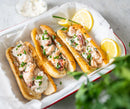 Three lobster rolls filled with creamy lobster salad and topped with chopped chives are served on a tray, accompanied by lemon wedges and a small bowl of sauce in the background.