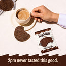 A hand holds a steaming cup of coffee next to a plate with a chocolate brownie wafer and an open package of RipVan Wafels, all set on a light-colored tablecloth.