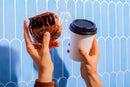 A person holds a cinnamon roll in a clear wrapper in one hand and a coffee cup with a black lid in the other, set against a light blue patterned background.