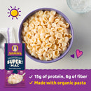 A purple bowl filled with creamy shell pasta sits on a light surface, next to a package of Annie's Super Mac, highlighting its protein and fiber content, with a glass of ice water nearby.