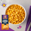 A bowl of creamy macaroni and cheese made with organic pasta sits on a light surface, accompanied by a box of Annie's Super Mac and a purple napkin, with playful graphics in the background.