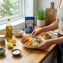 A person in a tank top adds fresh vegetables and couscous to a bowl on a wooden kitchen counter, with olive oil, spices, and a box of "Boost Couscous" visible in the background.