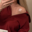 A person with long blonde hair is taking a selfie while wearing a maroon top, revealing a small bandage on their shoulder, and holding a phone in their hand.