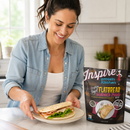 A woman smiles while placing a sandwich made with flatbread on a plate in a bright kitchen, with a package of Inspire protein kitchen flatbread mix visible nearby.