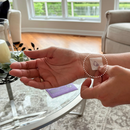 A person is applying a small adhesive patch to their wrist while seated in a cozy living room with large windows and a coffee table in the foreground.