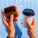 A person holds a packaged pastry in one hand and a coffee cup in the other, set against a light blue background with a decorative pattern.