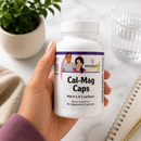 A hand holds a single capsule from a white supplement bottle labeled BariatricPal Cal-Mag Caps against a bright, clean background with promotional text and icons about bone health benefits.
