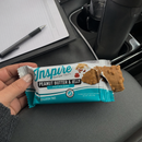 A hand holds an open package of Inspire Peanut Butter & Jelly protein bars, with a partially visible bar, resting on a car's console next to a pen and notepad.