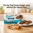 A package of Inspire Caramel Nut protein bars sits on a wooden table, surrounded by nuts and caramel pieces, with a bright kitchen setting in the background.
