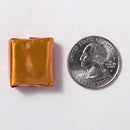 A small, shiny gold-wrapped candy square is placed next to a quarter on a white background, highlighting the candy's size in comparison to the coin.