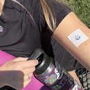 A person in a black shirt sits on a pink yoga mat, holding a water bottle with stickers, while a bandage with a blue symbol is visible on their arm.