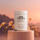 A white container labeled "HAIR VITAMINS. PLATINUM" with a gold logo sits on a light pedestal, surrounded by soft pink flowers against a serene sunset backdrop, promoting a dietary supplement for hair health.