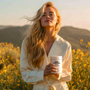 A woman with long, wavy hair stands in a field of yellow flowers, holding a container labeled "Hair Vitamins," with a serene expression against a sunset backdrop.