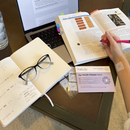 A hand holding a pink pen writes in a notebook beside a laptop and an open book, while a packet of Eye Health Vitamin Patches is displayed on a table.