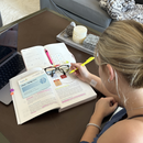 A young woman highlights text in an open book while studying at a table, with a laptop, glasses, and decorative items nearby, suggesting a focused learning environment.