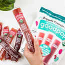 A hand holds a fruit punch popsicle made from 100% organic fruit juice, with several other popsicles and a colorful packaging labeled "goodpop" visible in the background.