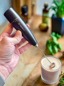 A hand holds a black milk frother above a glass of frothy beverage on a wooden countertop, surrounded by herbs and bottles in a cozy kitchen setting.